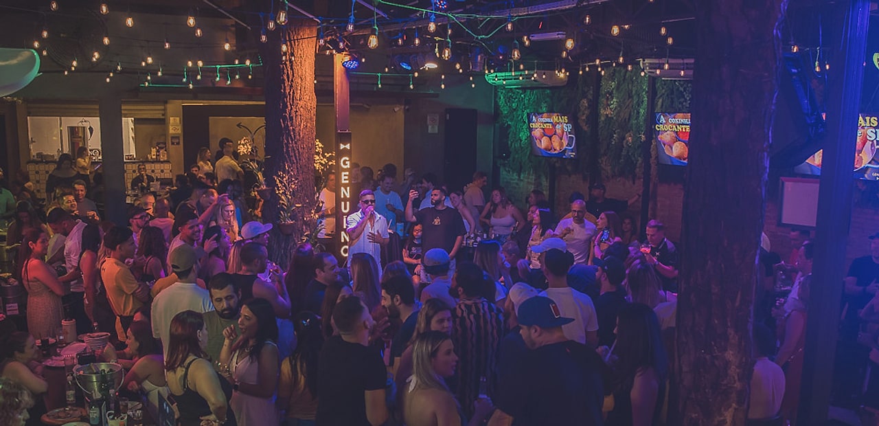 Salão do Genuíno Bar lotado numa noite de sábado na Vila Mariana, com varal de luzes, árvores e clientes de pé entre as mesas